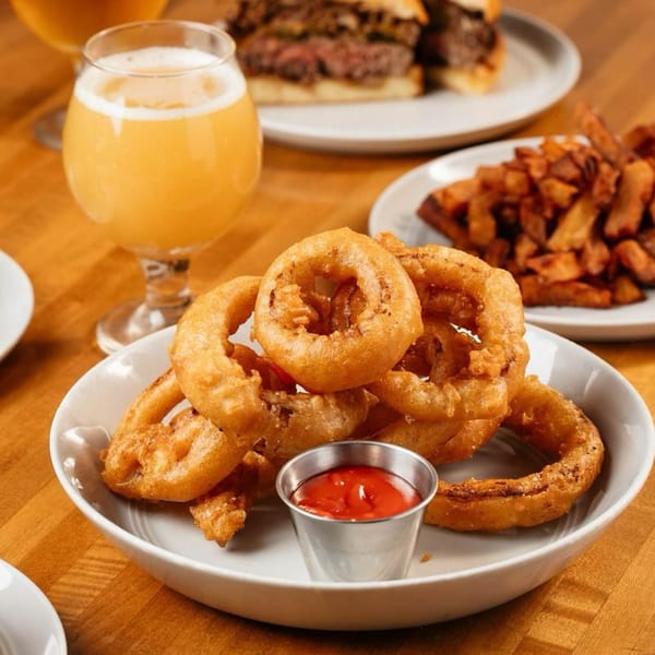 A plate of crispy onion rings with a side of ketchup, a burger, fries, and a beer.