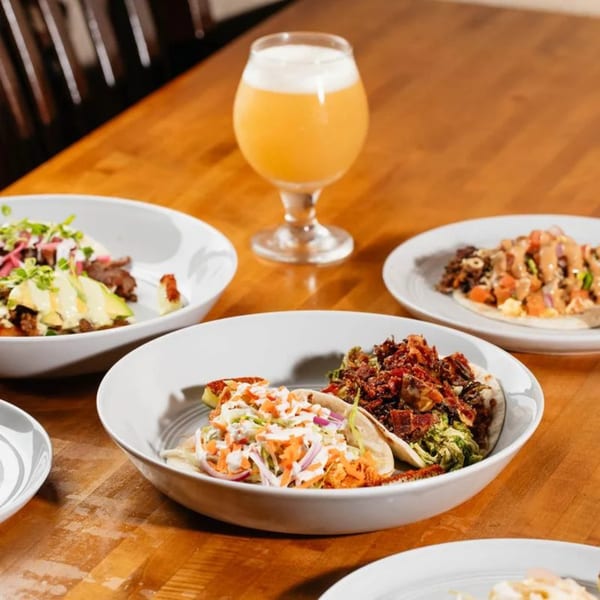 A wooden table displays several bowls of street tacos and a glass of beer.