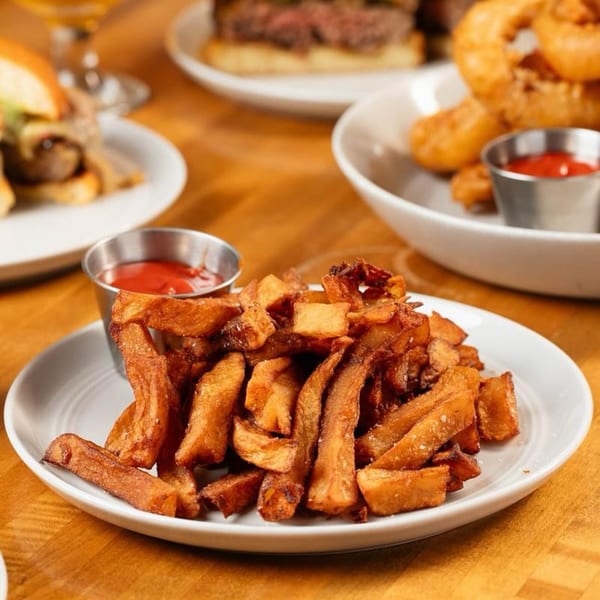 A plate of golden-brown, thick-cut fries with two small cups of ketchup on a wooden table.