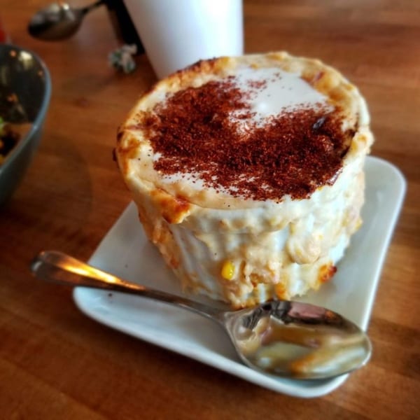 A cup of Mexican street corn, esquites, topped with white cream and chili powder, with a spoon.