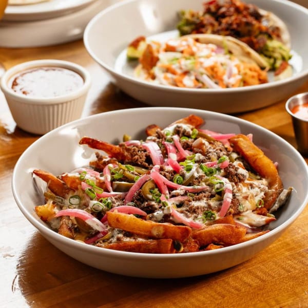 A bowl of loaded fries with ground beef, pickled onions, and sauce, with tacos in the background.