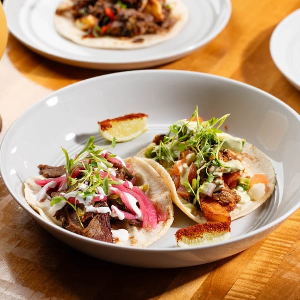 Two street tacos with various toppings, including meat, pickled onions, and microgreens, in a white bowl.